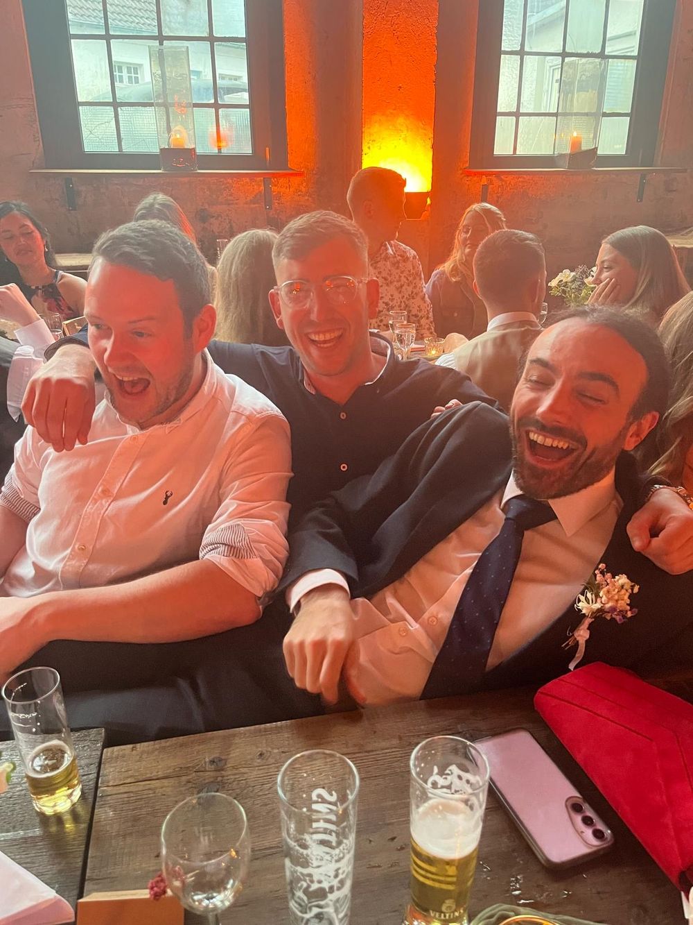 Three men laughing joyfully together at a wedding reception table with beer glasses in a warmly lit venue.
