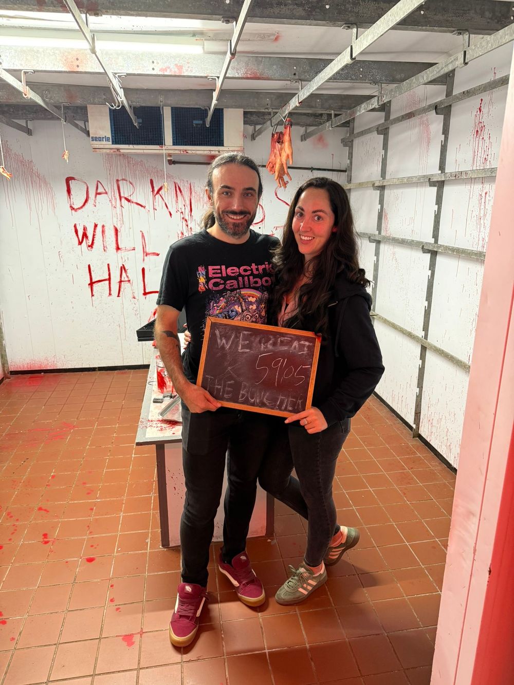 Couple posing with chalkboard sign in spooky Halloween escape room with red splatter walls.