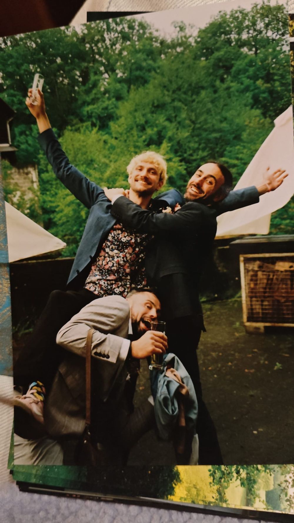Three men celebrating joyfully outdoors, two raising arms wide while one crouches between them.