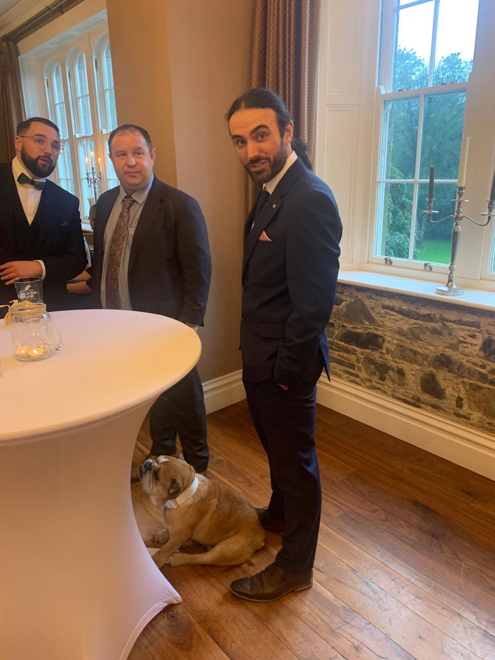 Men in suits at a formal event with a bulldog sitting on the floor beside a white cocktail table.