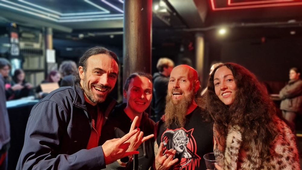 Four friends posing together at a dark music venue, making rock hand gestures and smiling at the camera.