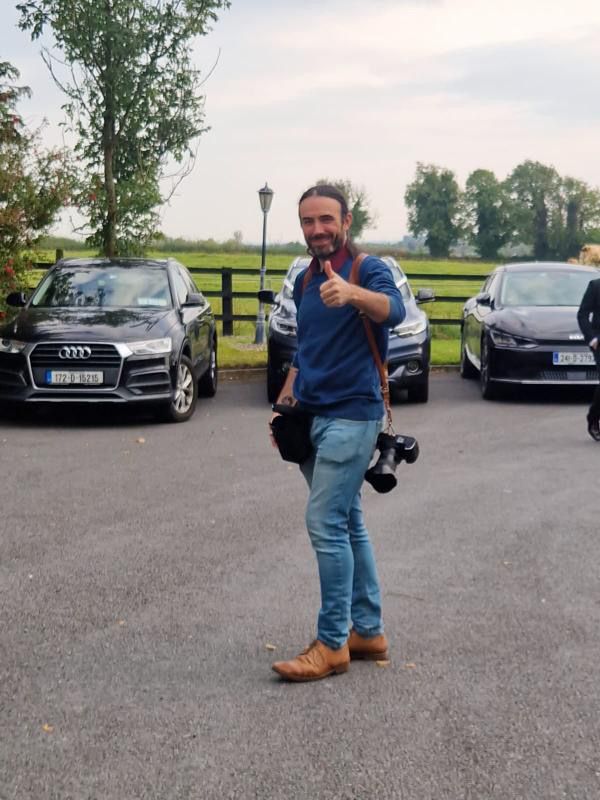 Man giving thumbs up in parking lot with cameras, wearing blue shirt and jeans, cars and green landscape behind.