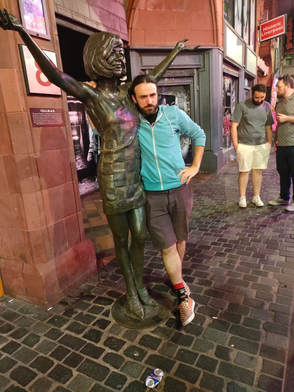 Bearded man posing beside a bronze street statue of a dancing figure in a city at night.