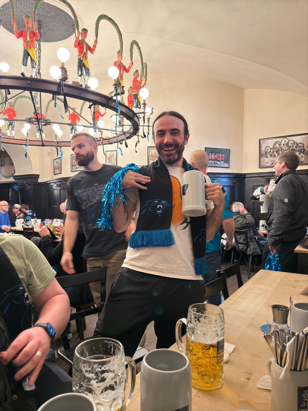 A smiling man holds a large beer stein and blue scarf in a festive German beer hall with decorative chandelier.