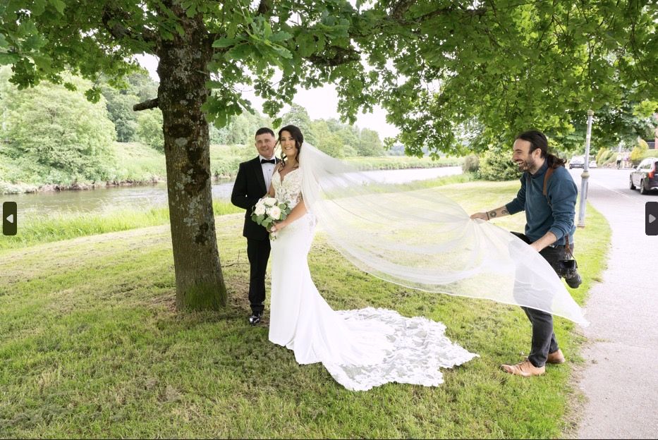 Wedding photographer captures bride and groom posing under a tree with a flowing veil outdoors.
