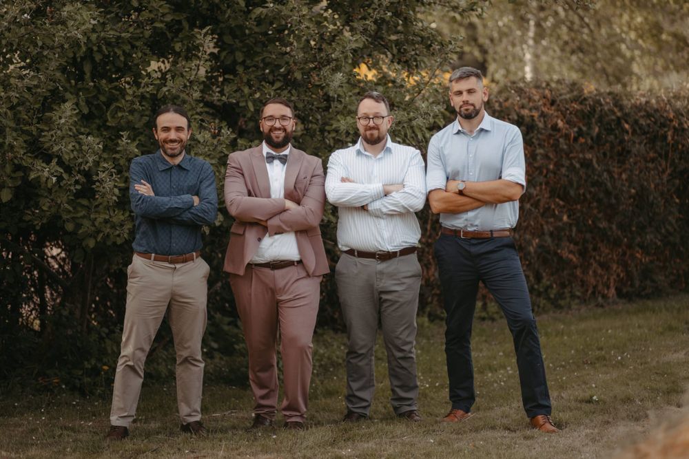 Four men in semi-formal attire stand smiling outdoors with arms crossed in front of lush green trees.