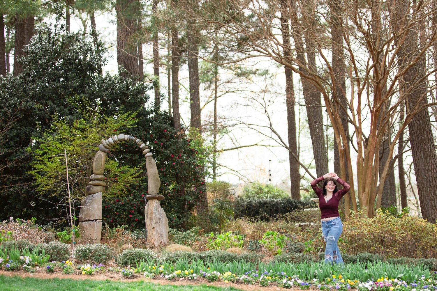 Woman posing in a lush garden with a stone arch and tall trees in the background at WRAL Azalea Garden