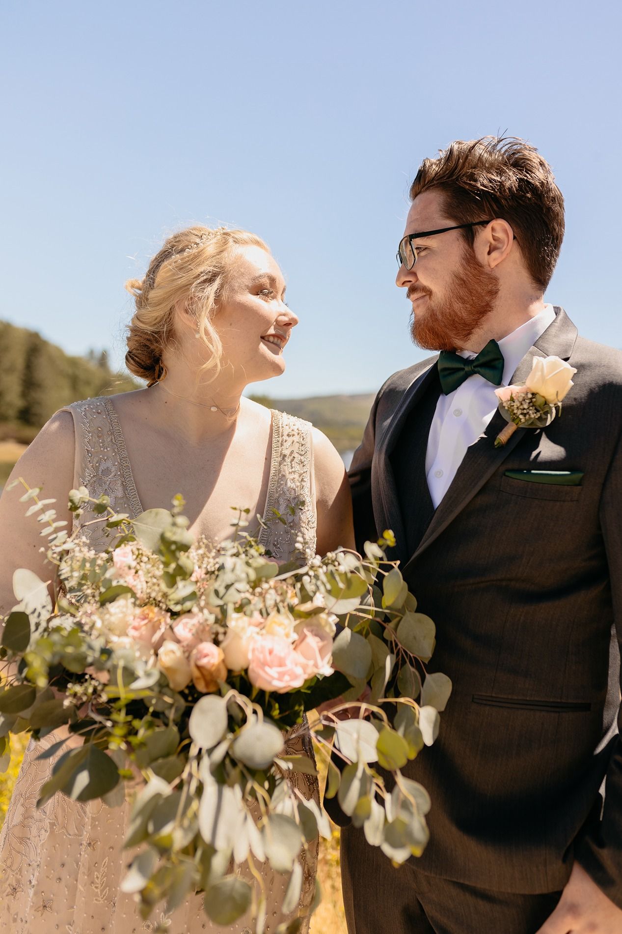 Bride and groom smiling at each other outdoors, holding a lush bouquet of pink roses and eucalyptus on a sunny day at Lake Cuyamaca, Julian.