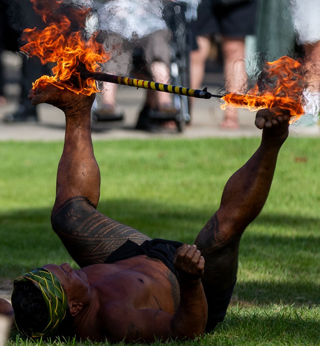 Muscular tattooed fire dancer lying on grass, balancing a flaming staff with his feet during an outdoor performance.