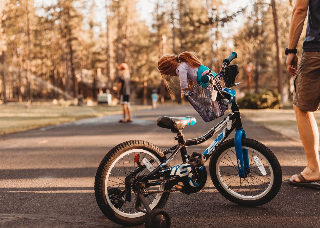 Child in helmet stands beside a small blue bicycle on a paved path surrounded by tall trees in a park.