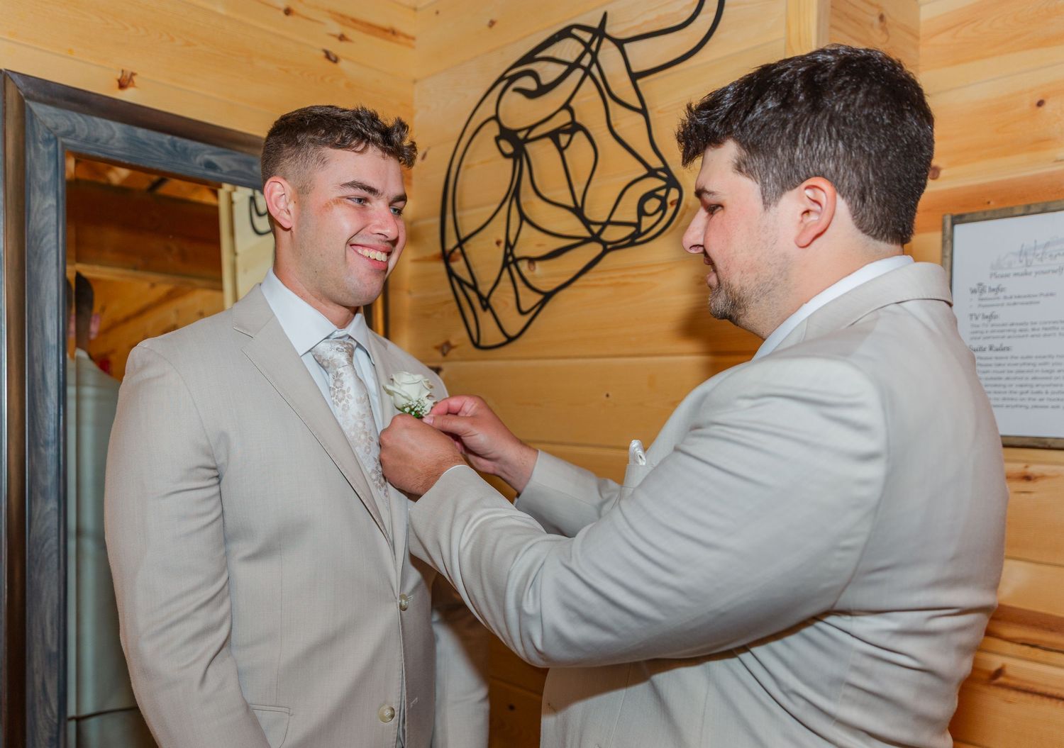 Two men in tan suits at a wedding, one pinning a boutonniere on the other inside a rustic wood-paneled venue.