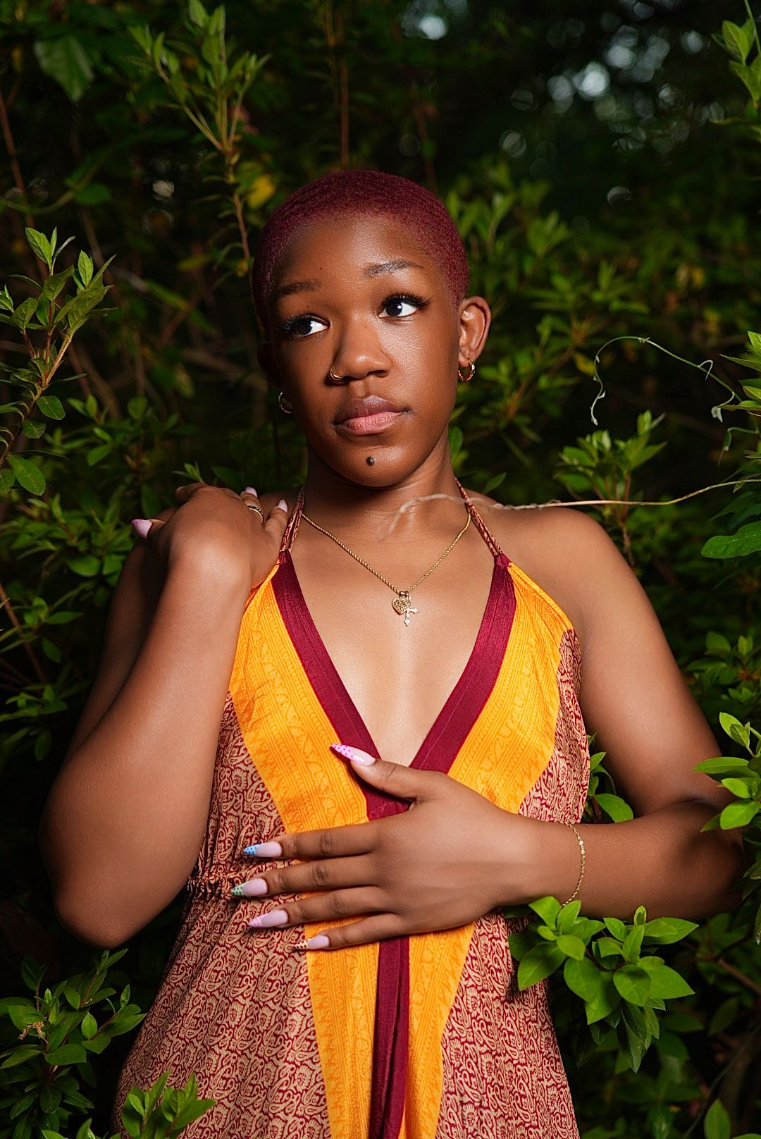 A young woman with short dyed red hair wears an orange and maroon halter dress and poses among green leaves in a forest.