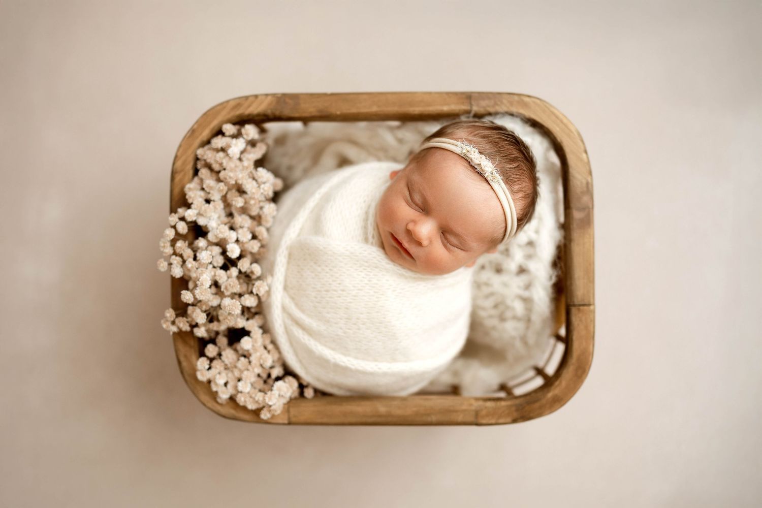 Newborn baby swaddled in white wrap sleeping peacefully in a wooden basket with delicate white flowers.