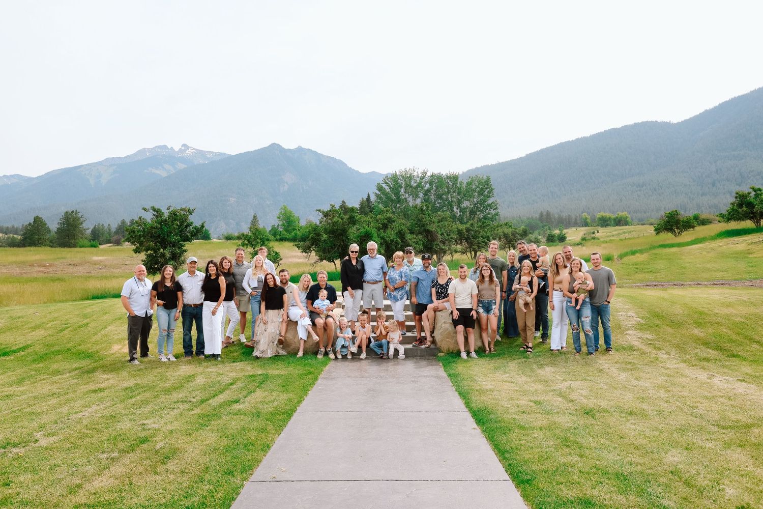 Large family group portrait outdoors on green grass surrounded by tall trees on a sunny day.