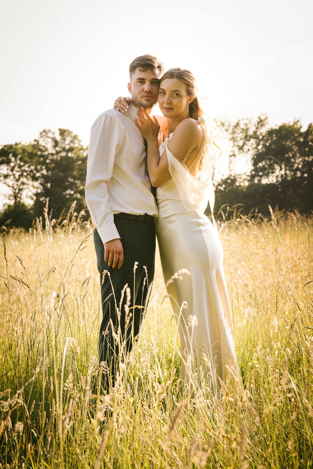 Wedding couple sharing a romantic kiss at golden hour sunset in a green field, bride in lace gown lifted by groom at Oak Barn, Frame Farm.