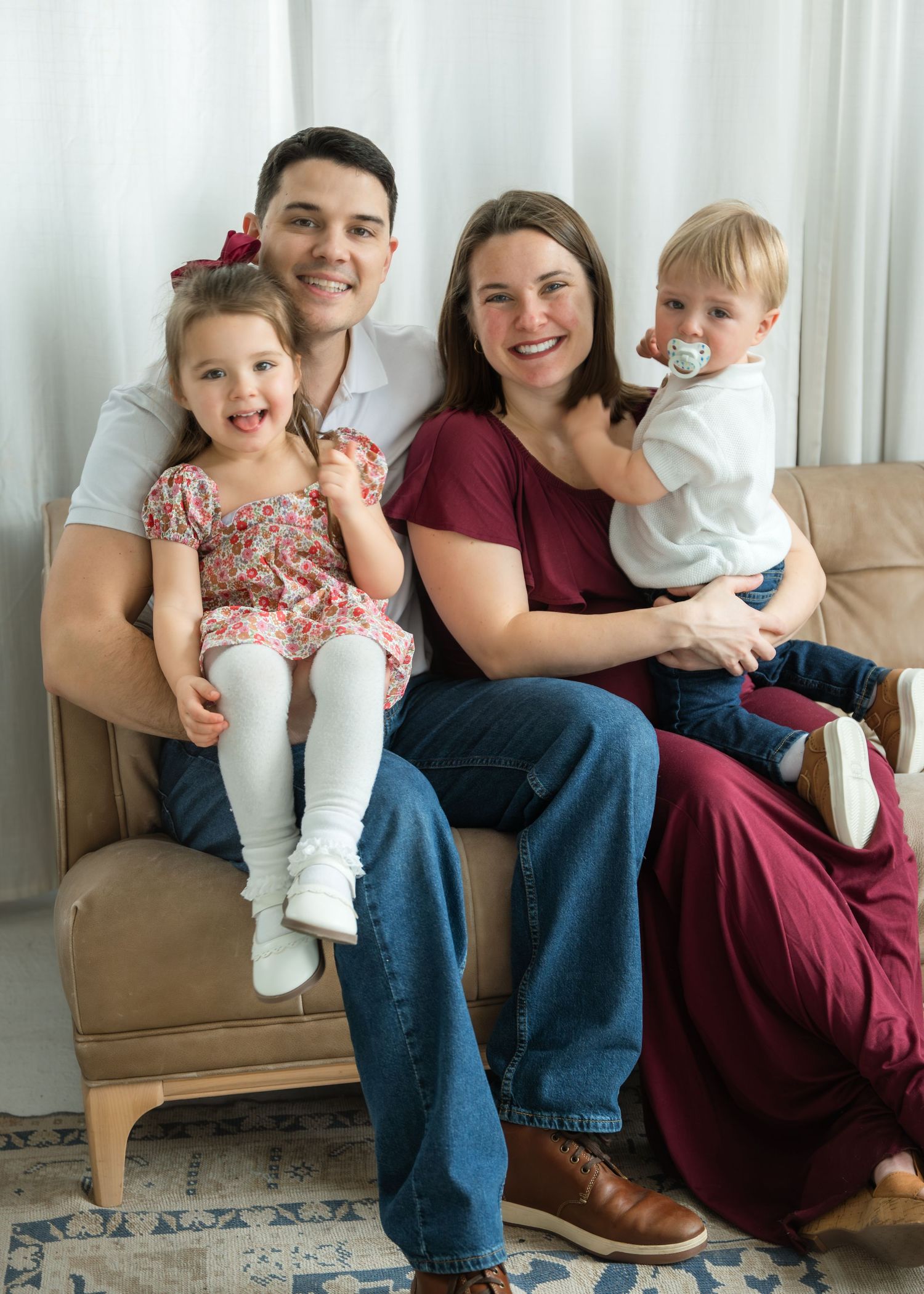 A happy family of four poses on a couch, with parents holding a young girl in a floral dress and a toddler boy with a pacifier.