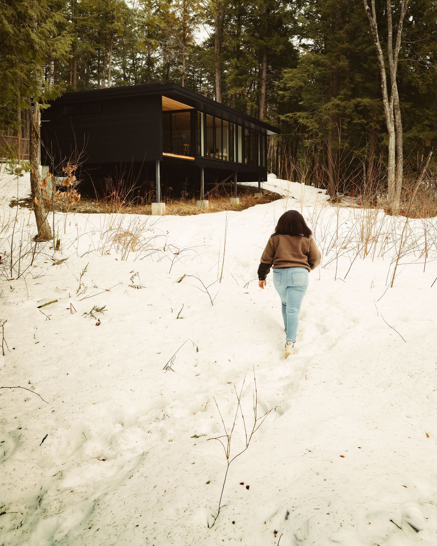 Woman walking through snowy path toward a modern dark cabin surrounded by winter trees.