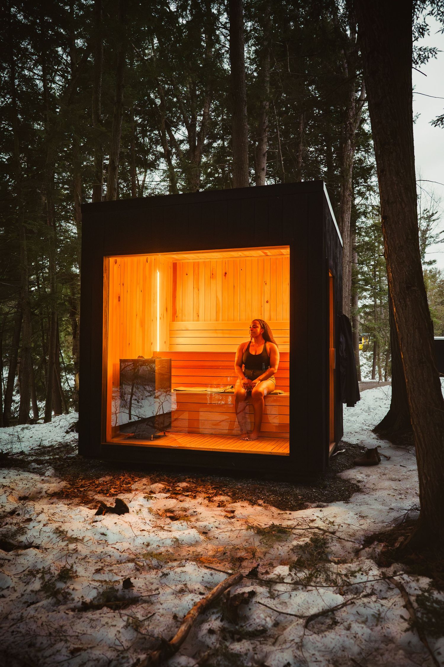 Woman relaxing inside a glowing cedar sauna cabin surrounded by snow-dusted forest trees in winter.
