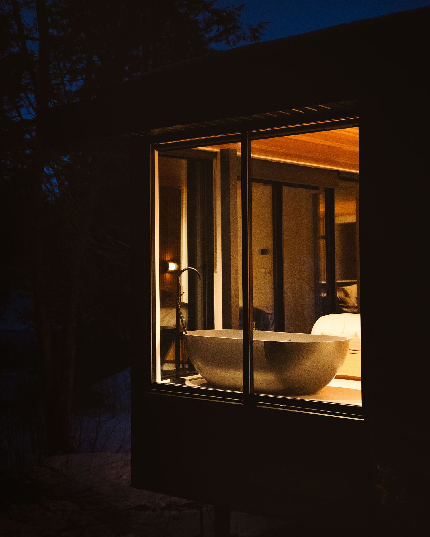 A glowing bathroom window at dusk reveals a freestanding soaking tub inside a modern luxury villa at night.