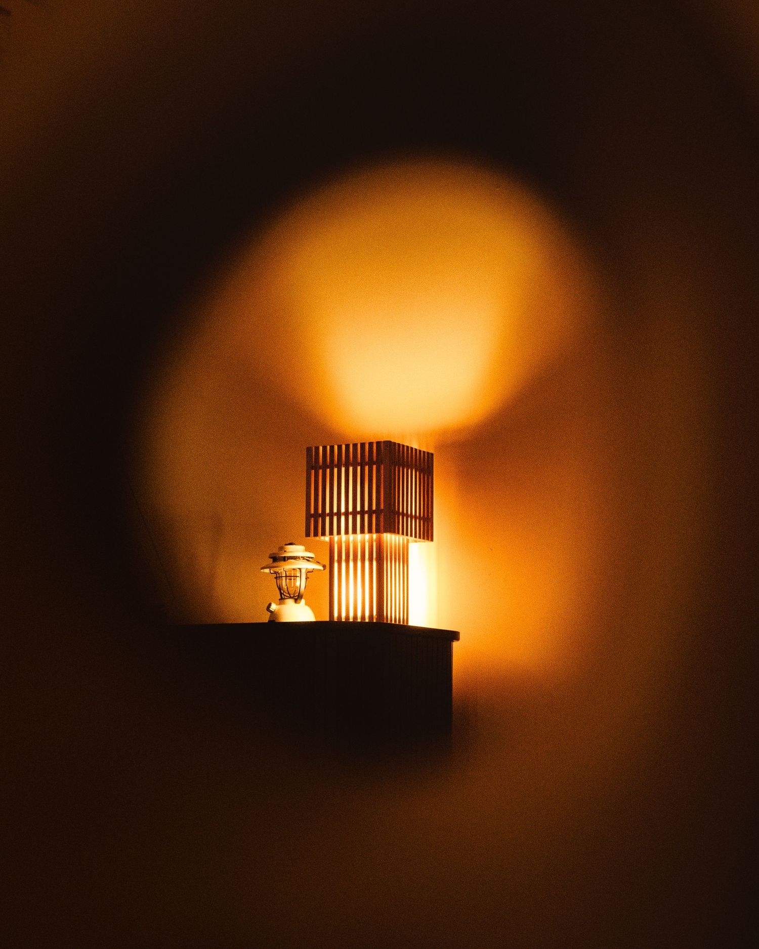 A warm glowing wooden lantern on a wall shelf casting a soft orange light circle on the wall.