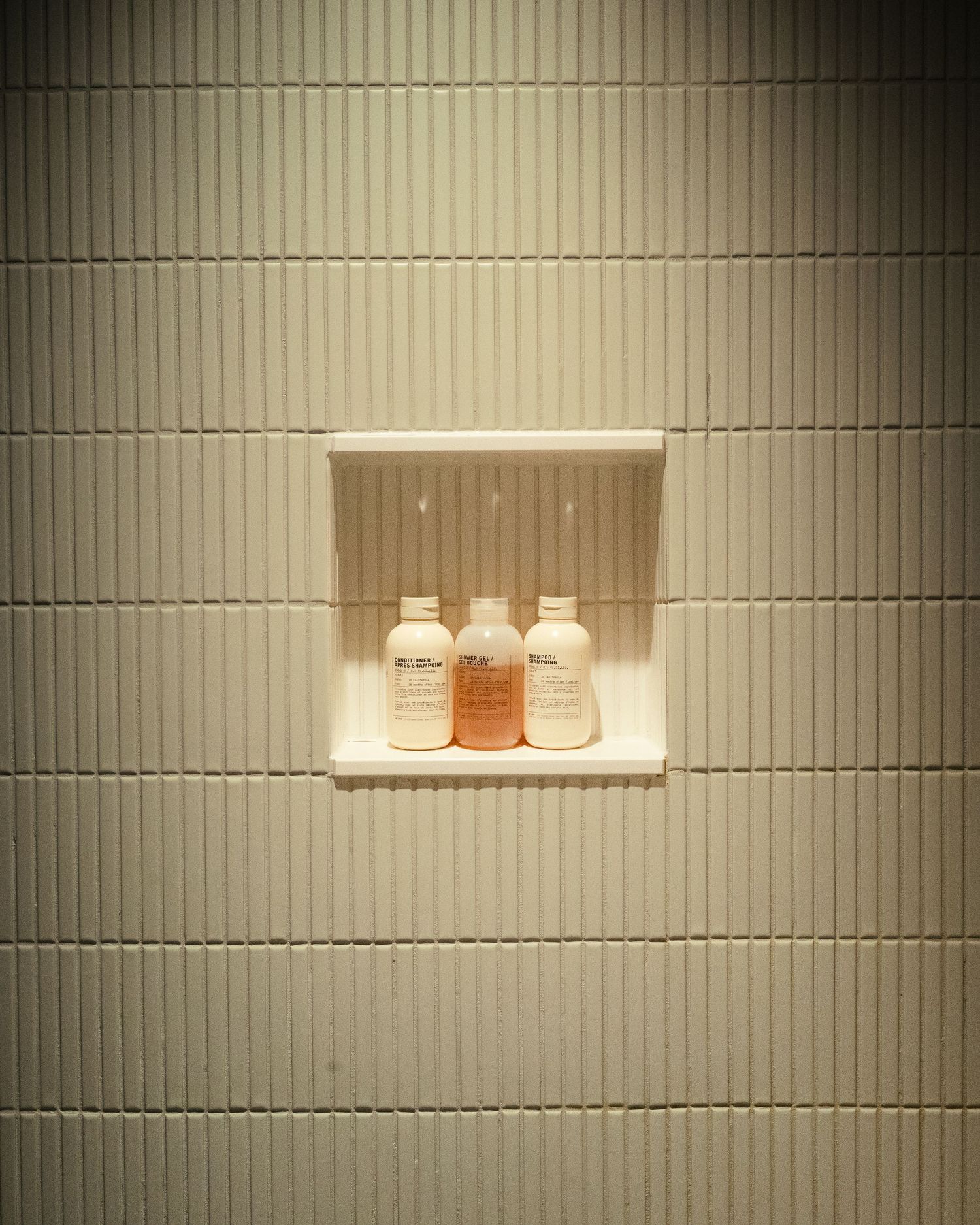 Three hotel toiletry bottles on a recessed white tile shower niche shelf in a modern bathroom.