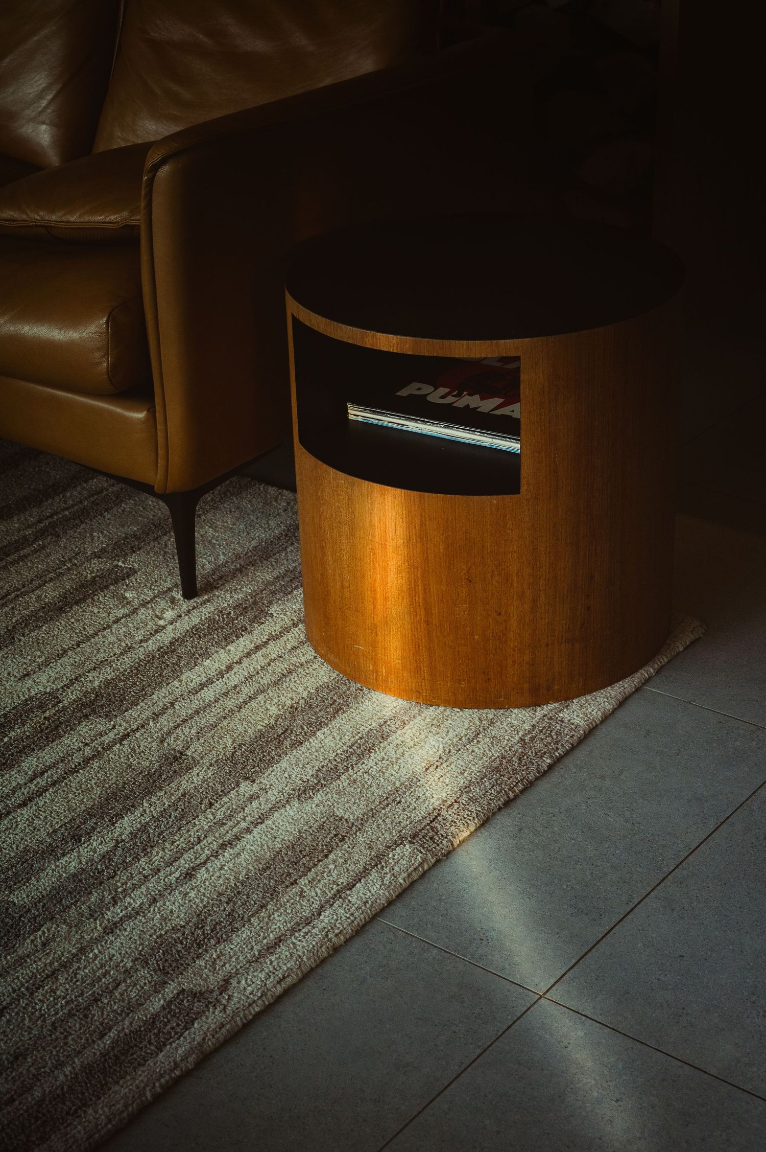 A warm-toned leather armchair beside a cylindrical wooden side table with a Puma item on a textured rug in dim light.