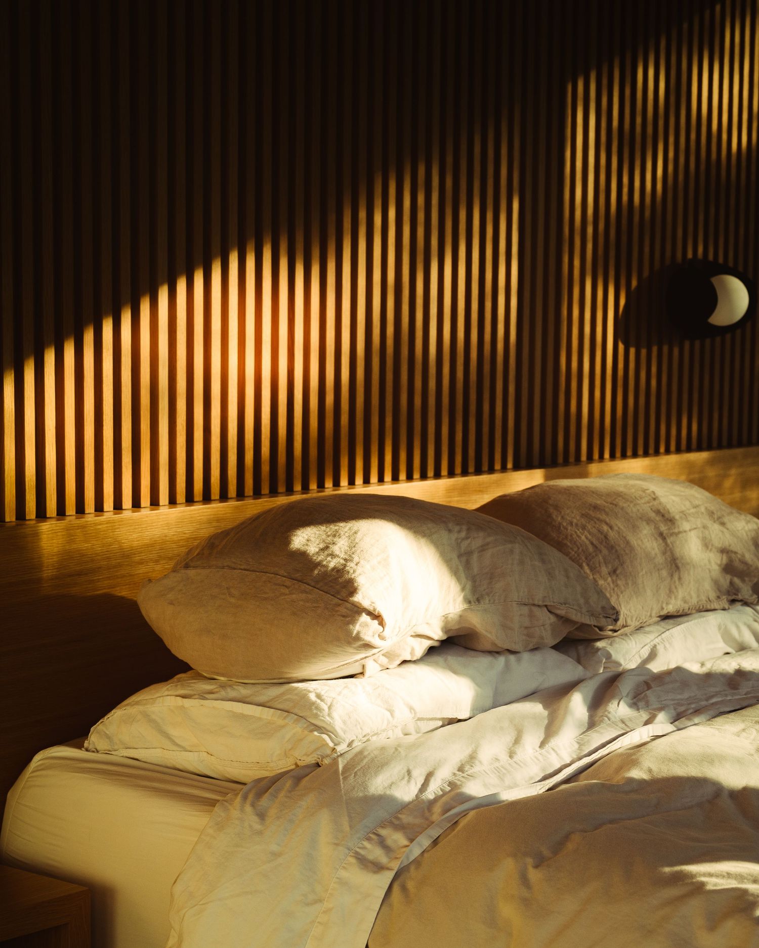 Warm golden sunlight casts shadows on a slatted wood headboard above a neatly made bed with cream linen pillows.