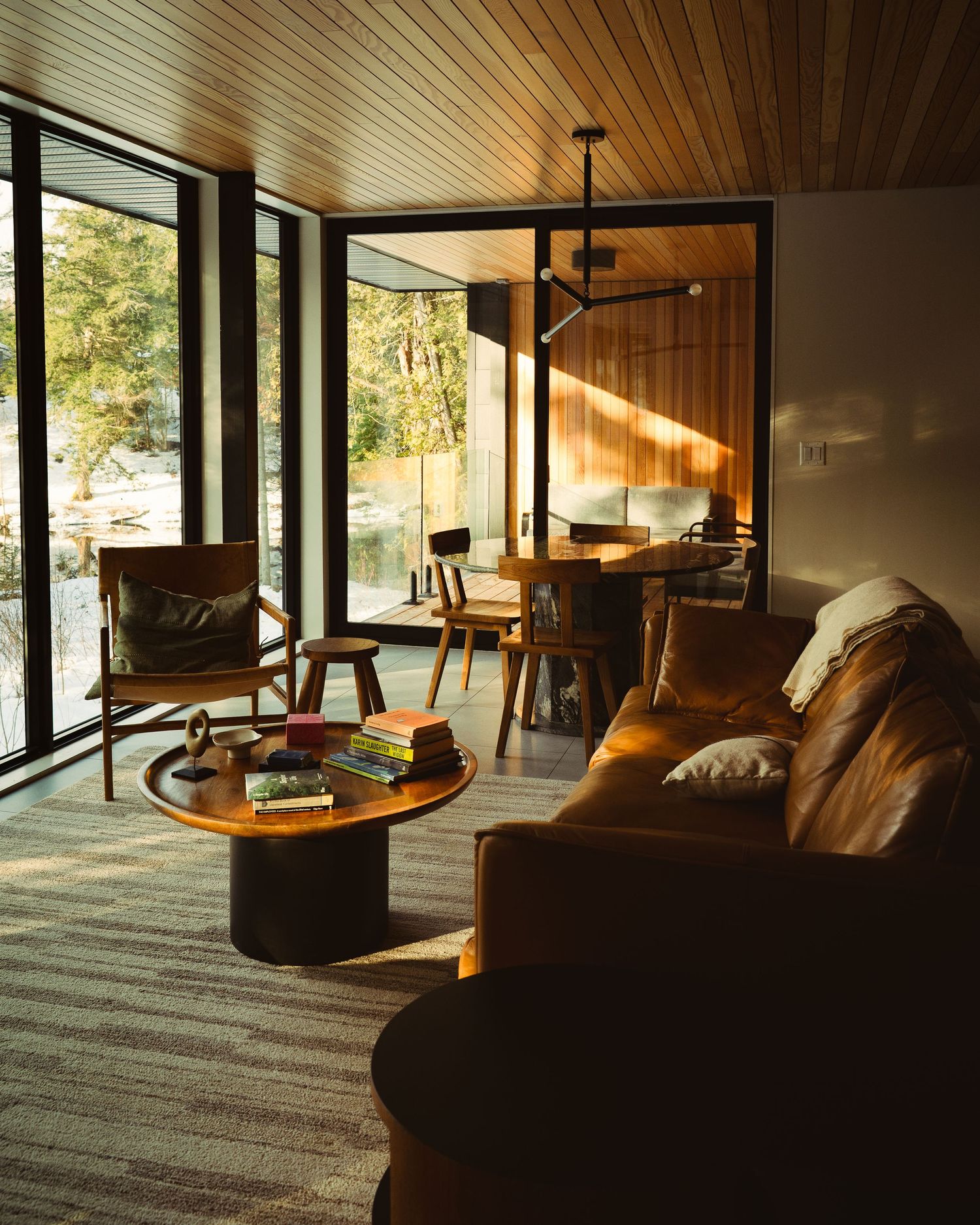 Warm mid-century modern living room with leather sofa, wood ceiling, large windows, and golden sunlight streaming through glass doors.