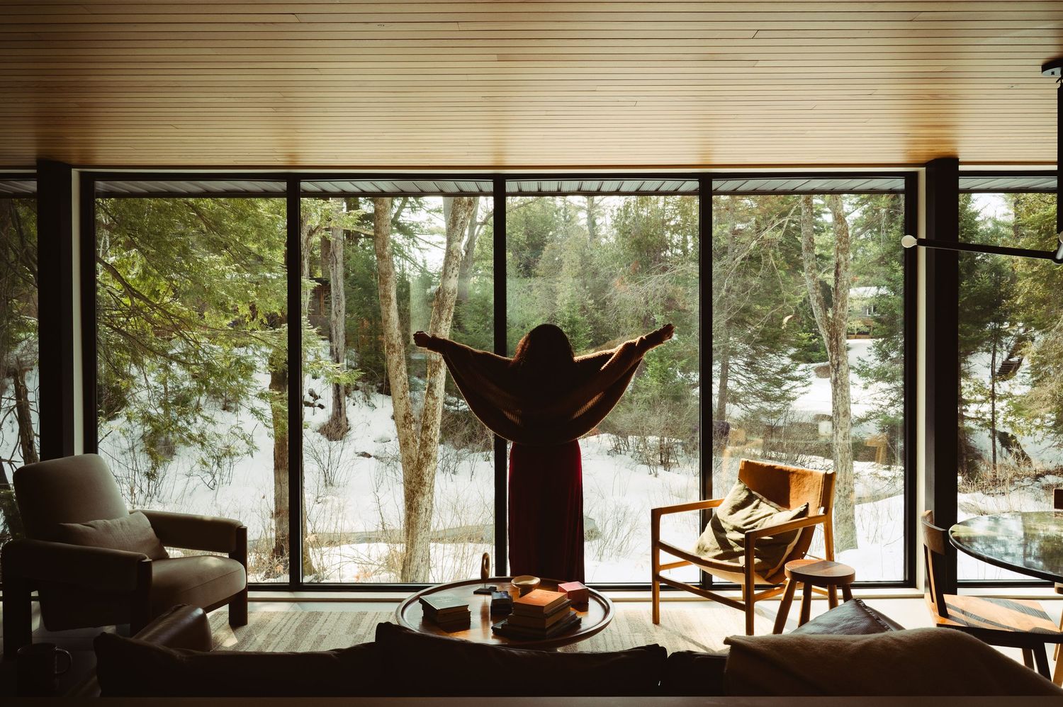 Person standing with arms outstretched before large windows overlooking a snowy forest in a cozy modern cabin interior.