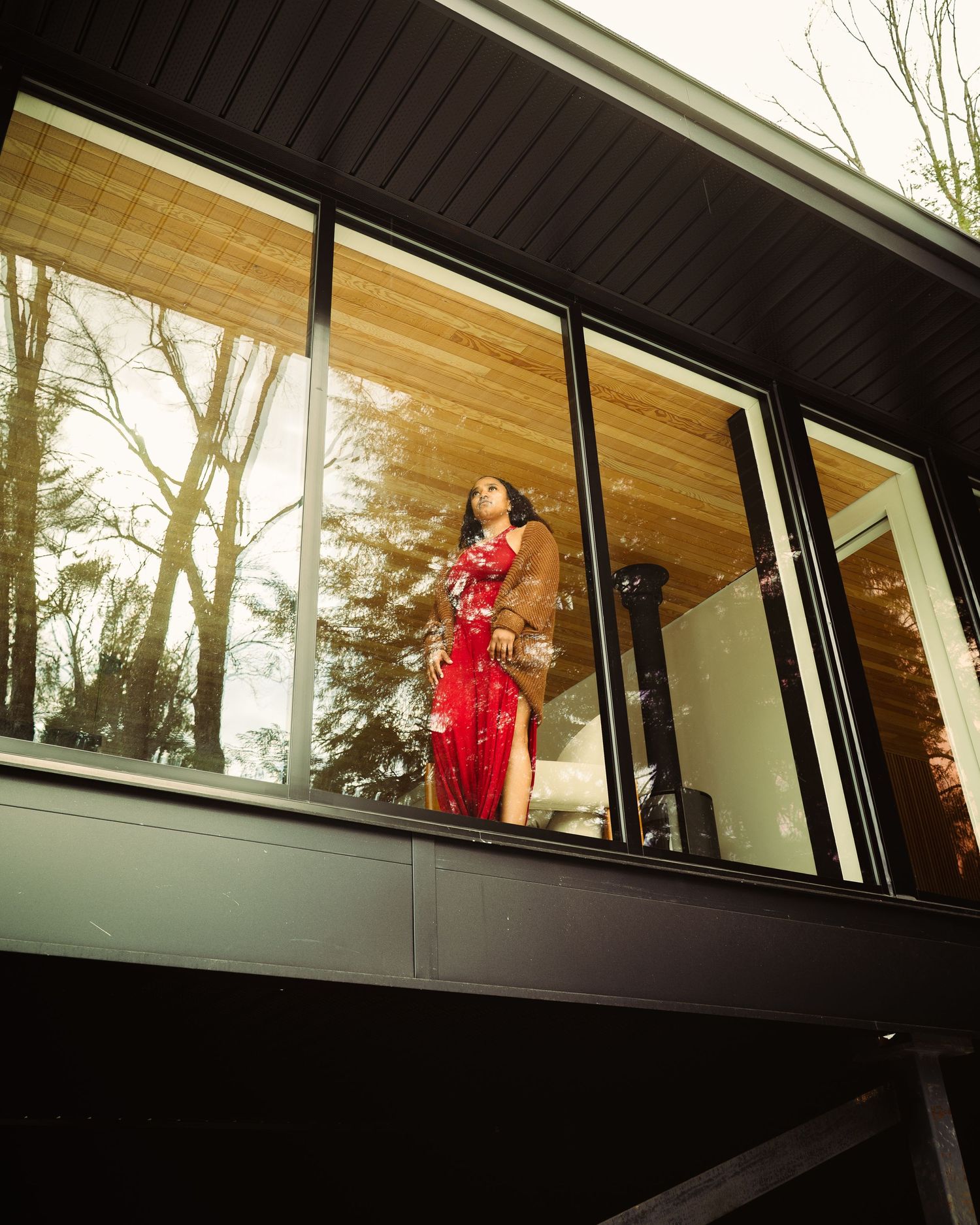 Woman in red dress and brown jacket stands on modern black-framed glass balcony surrounded by trees.