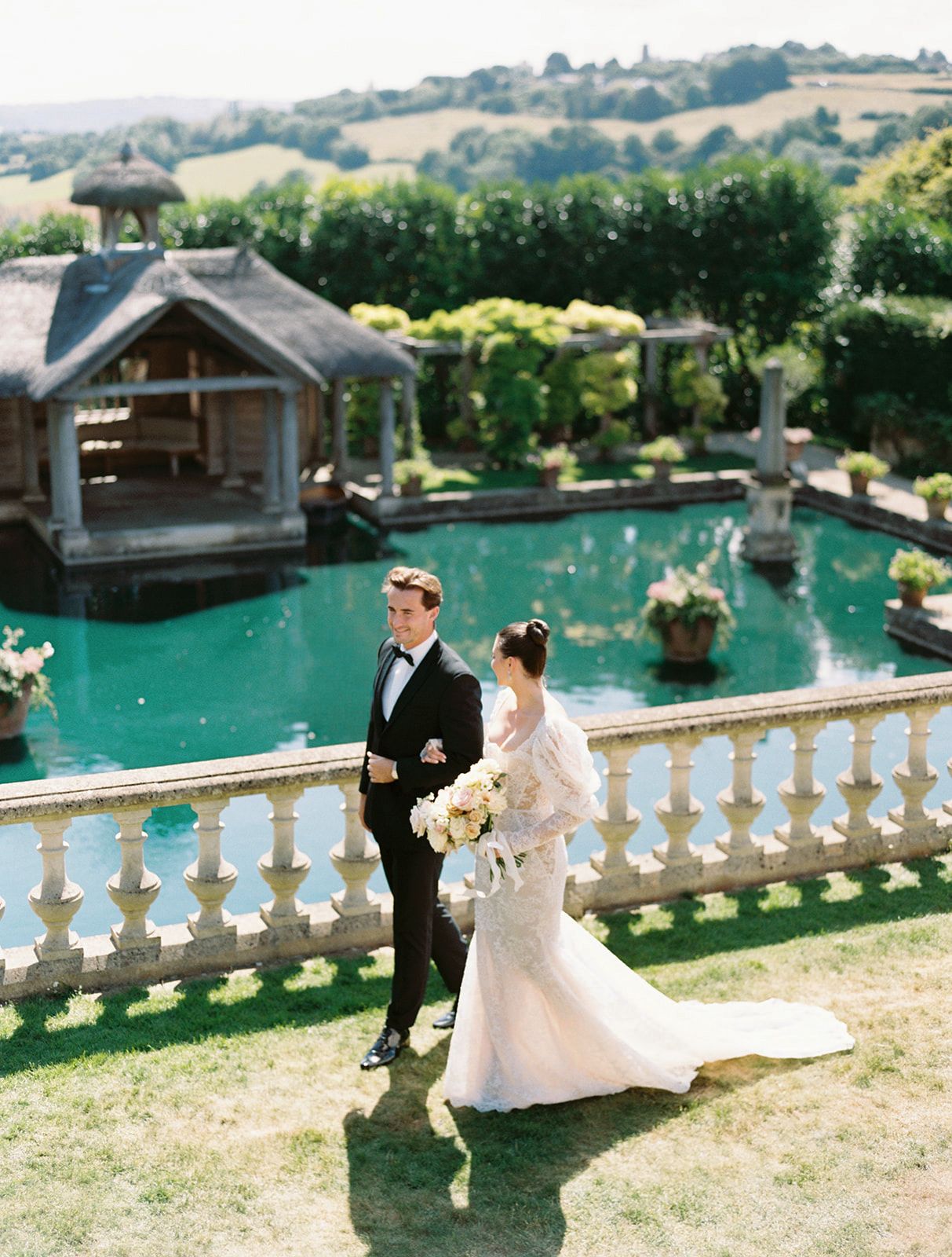 Bride and groom pose on stone balcony overlooking turquoise pond at elegant outdoor wedding venue.