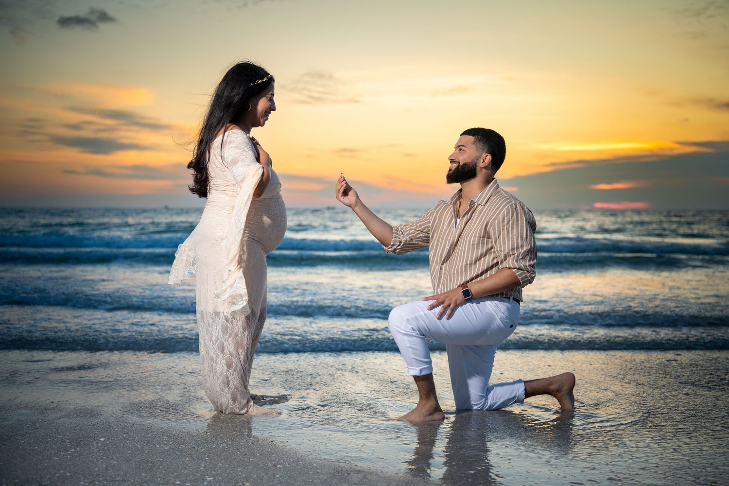 Man proposes to pregnant woman on beach at sunset, both dressed in white, waves and colorful sky in background.