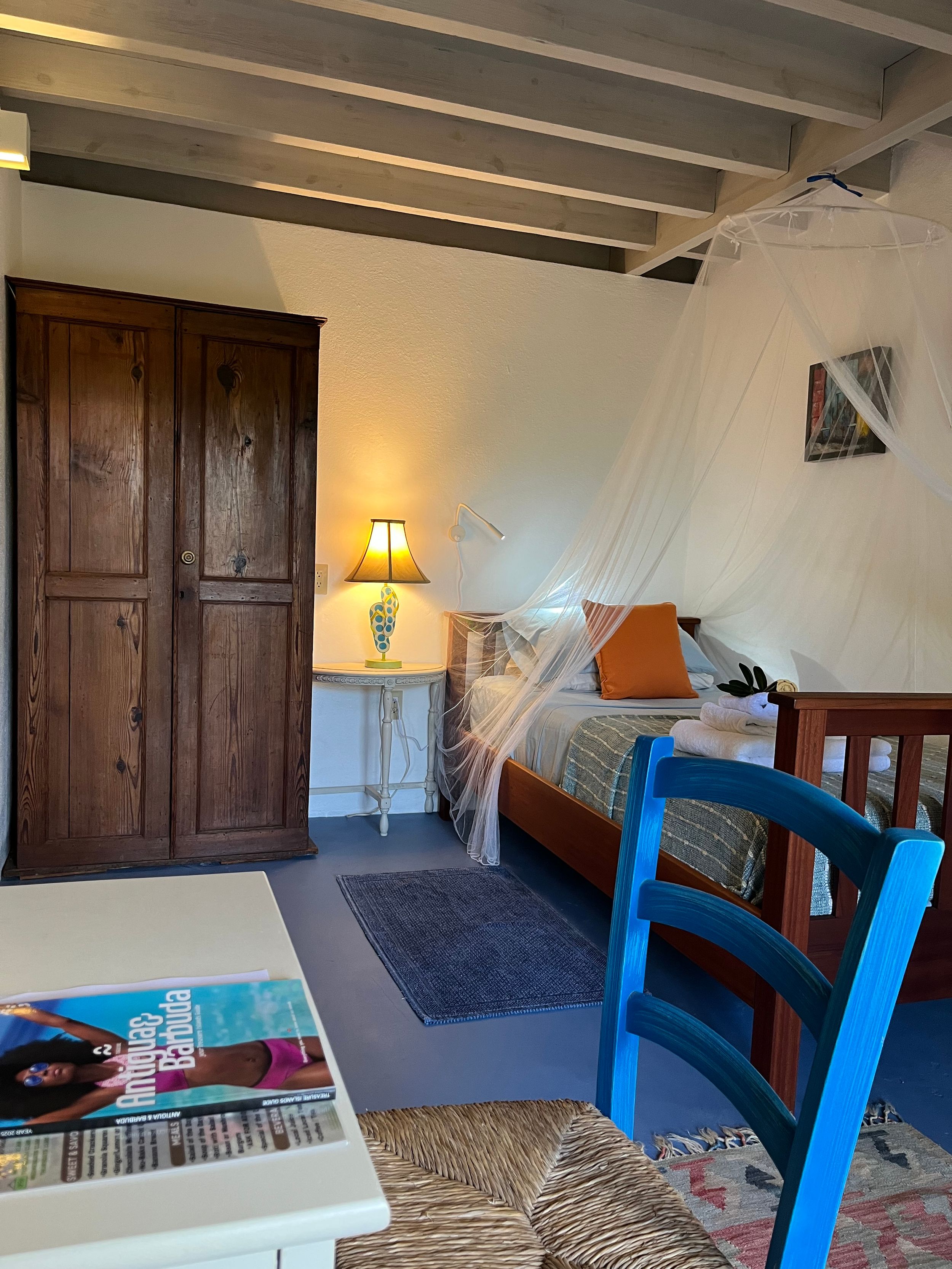 Cozy rustic bedroom with wooden wardrobe, mosquito net canopy bed, blue chair, and warm lamp lighting.