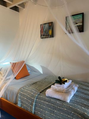 Cozy bedroom with canopy mosquito net over a bed with gray bedding, orange pillow, folded towels, and tropical artwork.