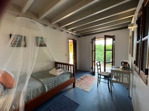Cozy guesthouse room with mosquito net canopy bed, wooden ceiling, tiled floor, small desk, and open doors to a lush garden.