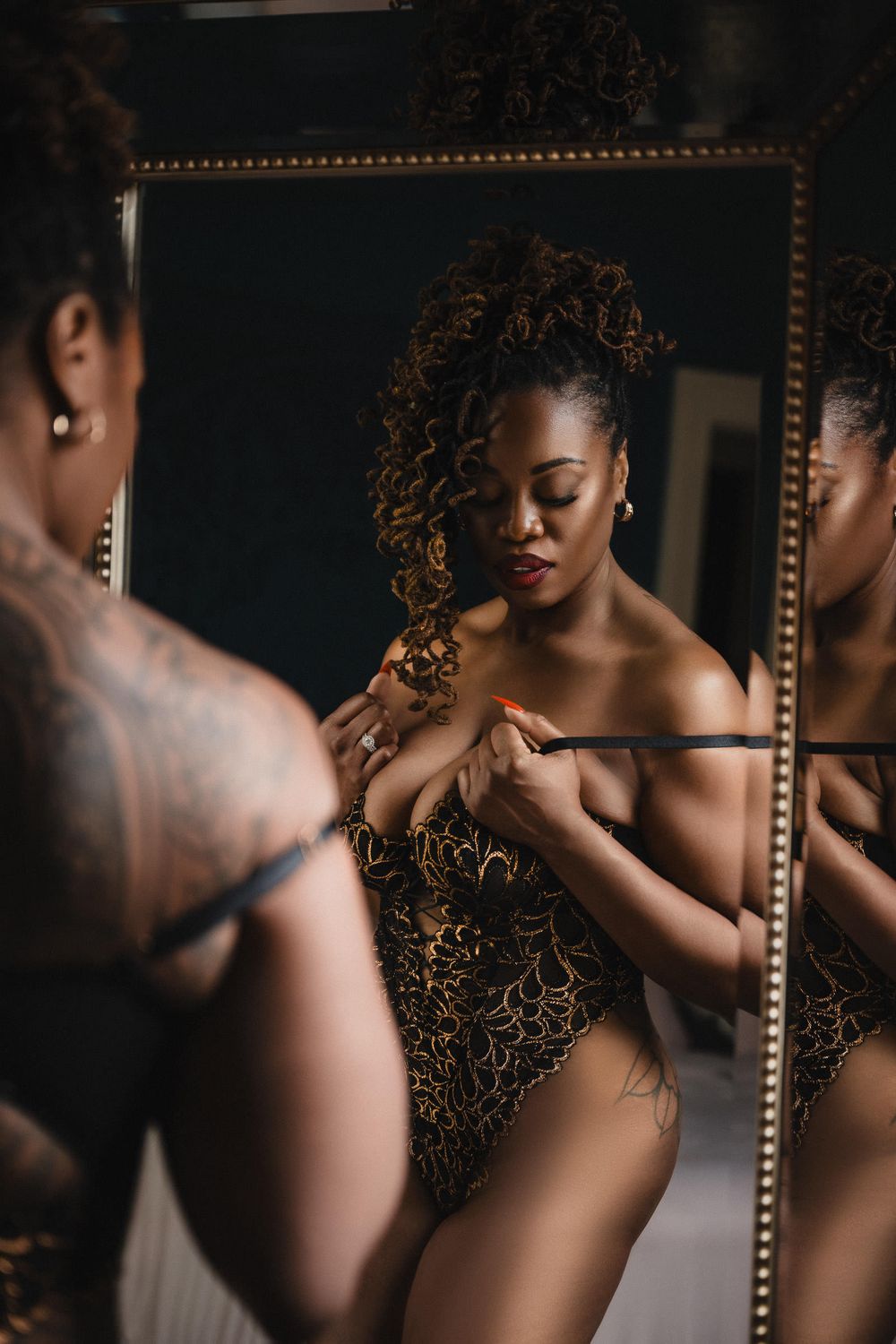 New Orleans Boudoir Photography - A woman in gold lace lingerie adjusts her outfit while gazing into an ornate gold-framed mirror.