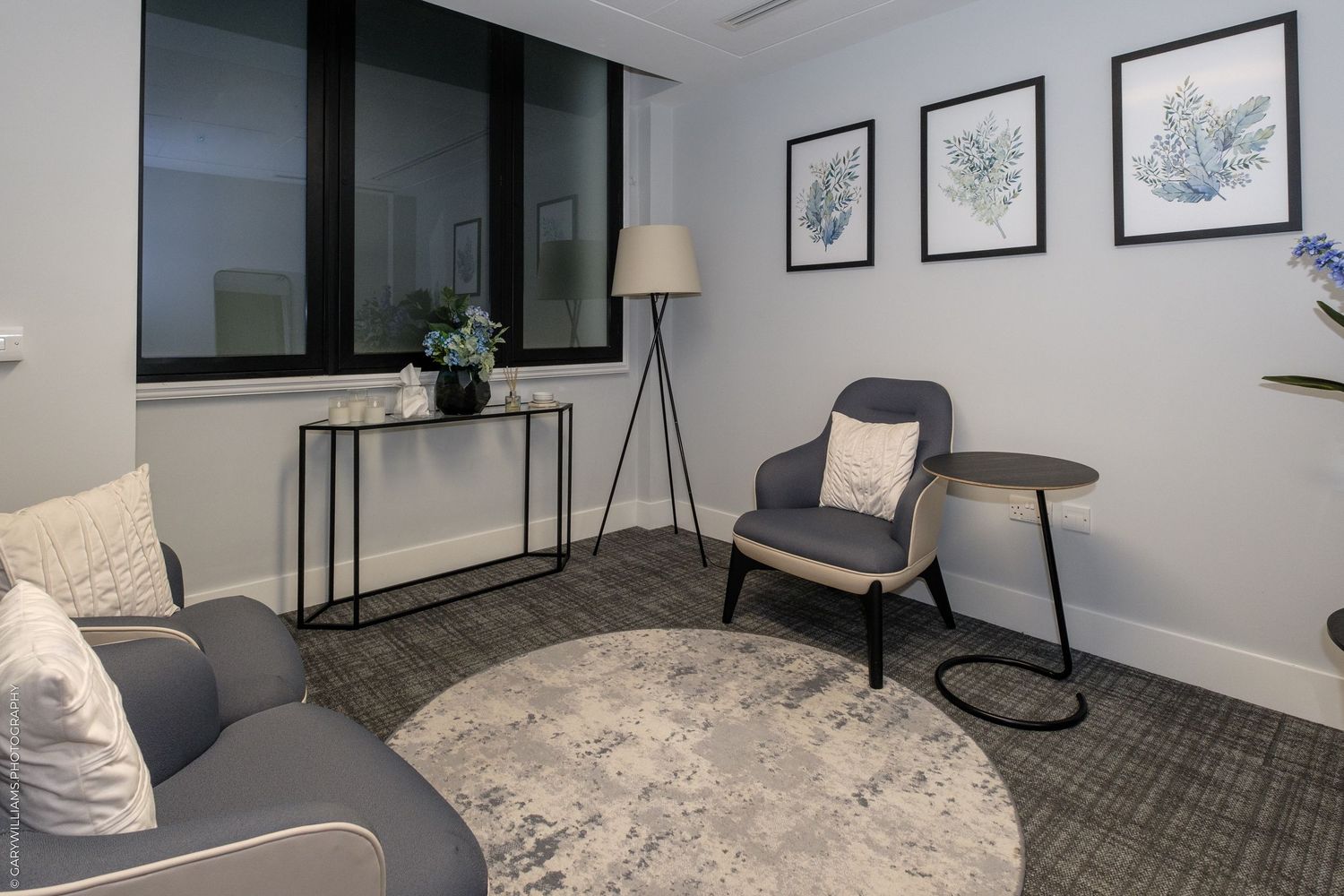 Modern living room with grey armchair, round rug, floor lamp, console table, and botanical wall art prints.