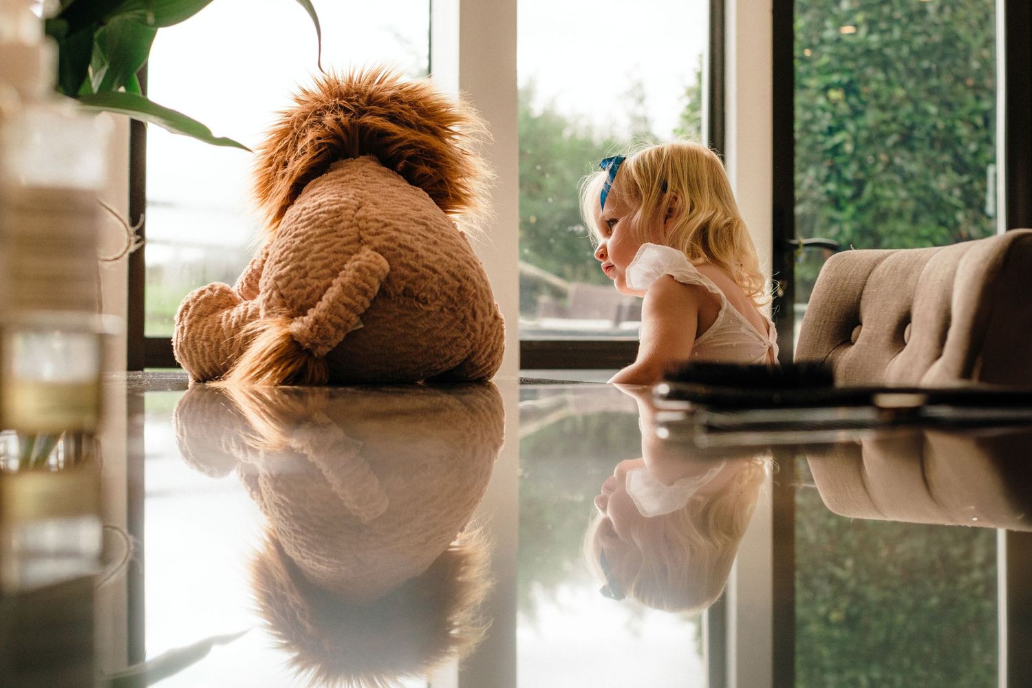 A toddler sits beside a large teddy bear on a reflective floor near a sunlit window indoors.