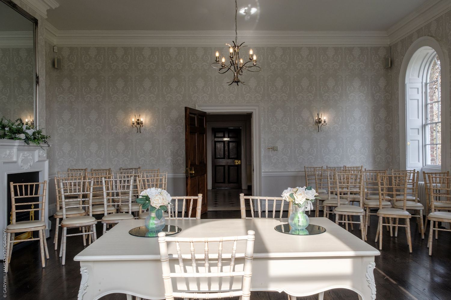 Elegant wedding ceremony room with gold Chiavari chairs, white tables with floral centerpieces, chandelier, and ornate wallpaper.