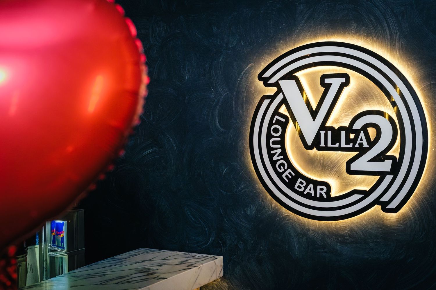 Red balloon beside illuminated Villa 2 Lounge Bar circular logo sign on dark textured wall.
