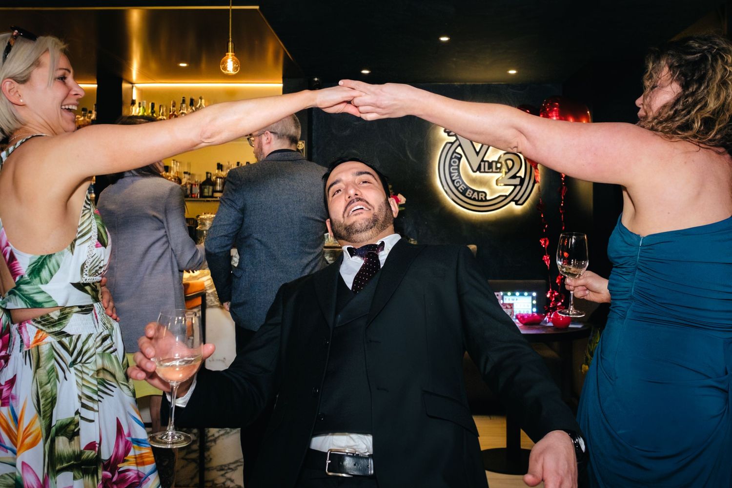 A man in a tuxedo dances joyfully as two women raise their arms over him at a lively wedding reception bar.