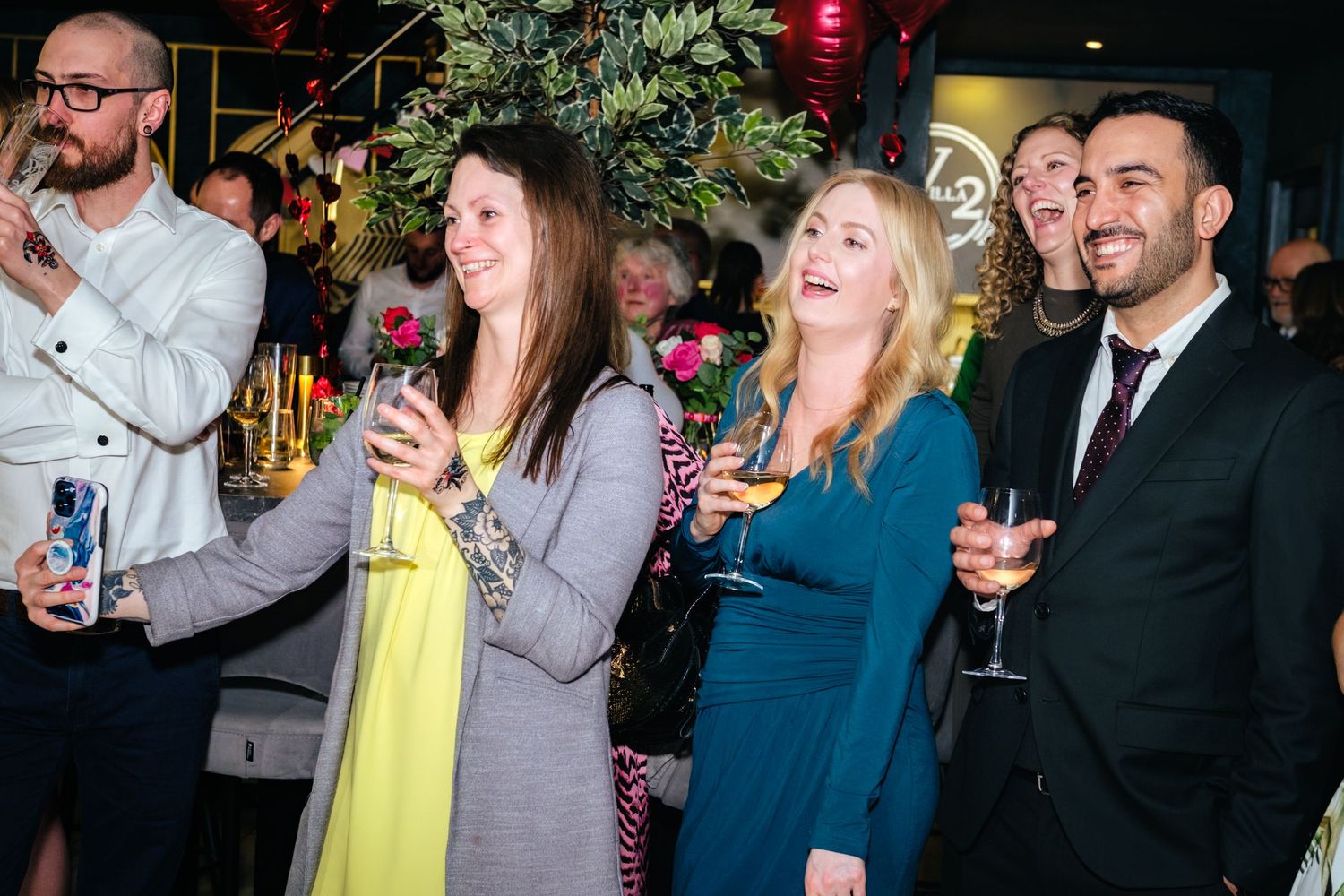 Group of smiling people holding drinks at a lively indoor party event with colorful floral decorations.