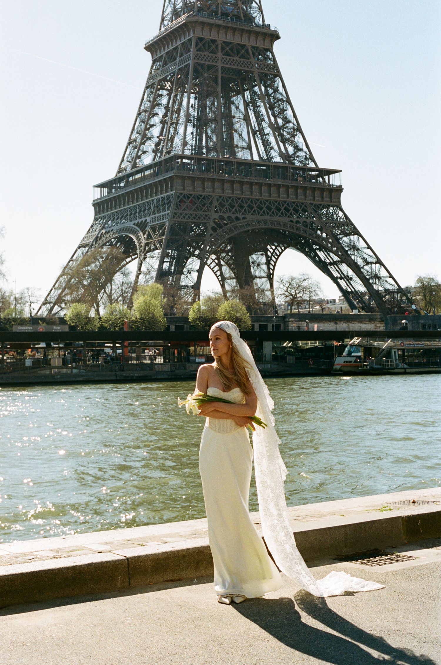 Bride in white gown and veil poses by the Seine River with the Eiffel Tower in Paris, France backdrop.