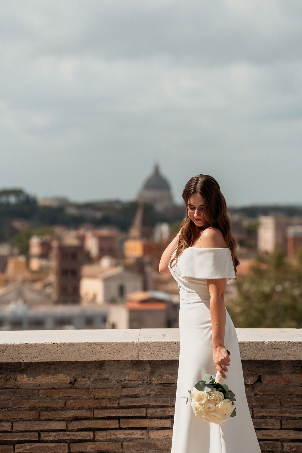 Elegant bride in off-shoulder white gown holding bouquet with Rome cityscape and St Peters dome behind her.