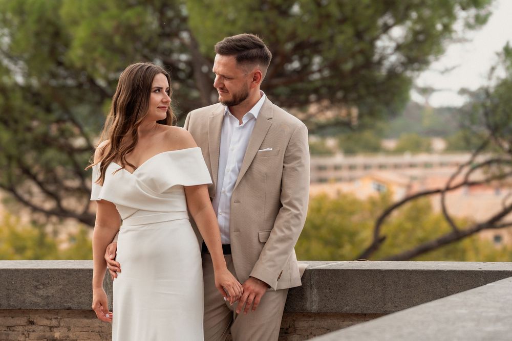 Elegant couple in wedding attire holding hands outdoors, woman in white off-shoulder gown, man in beige suit.