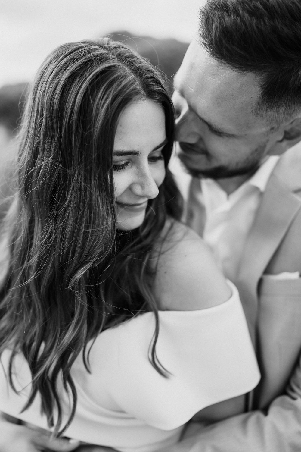 Black and white photo of a couple embracing, woman in off-shoulder top smiling softly as man leans in close.
