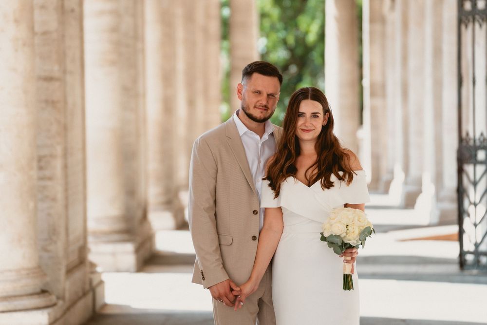 Couple in wedding attire hold hands among classical columns in Rome.