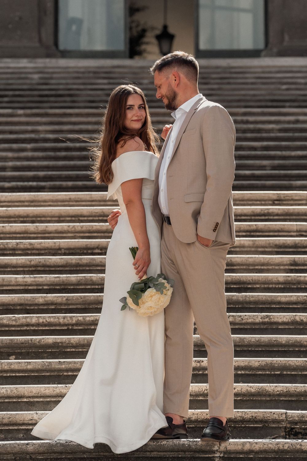 Elegant bride in white off-shoulder gown and groom in beige suit pose on stone steps holding a white bouquet.