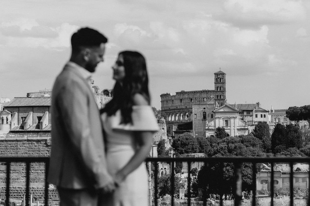 Black and white photo of a couple holding hands with the Roman Colosseum and ancient ruins in the background.