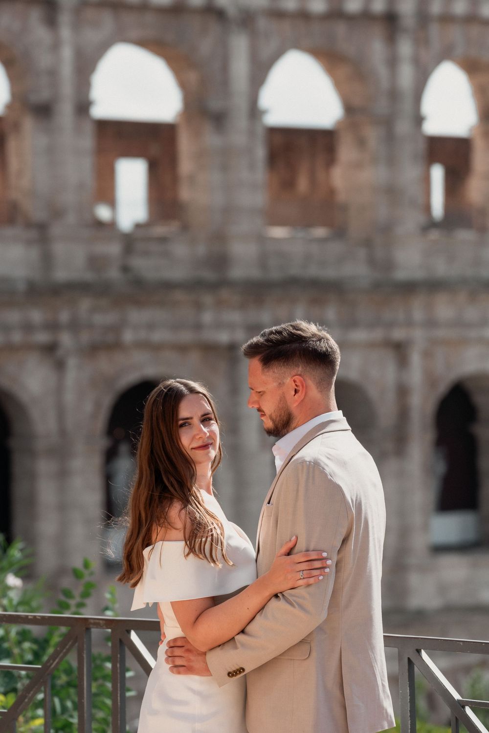 Couple embracing romantically in front of the ancient Colosseum in Rome, woman in white dress, man in beige suit.